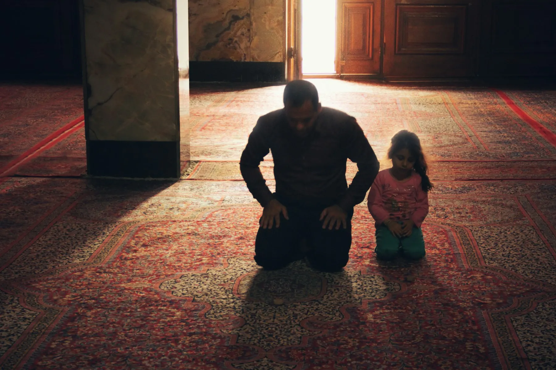 Un padre y su hija pequeña rezan juntos sobre la alfombra de una mezquita, vistos de espaldas.