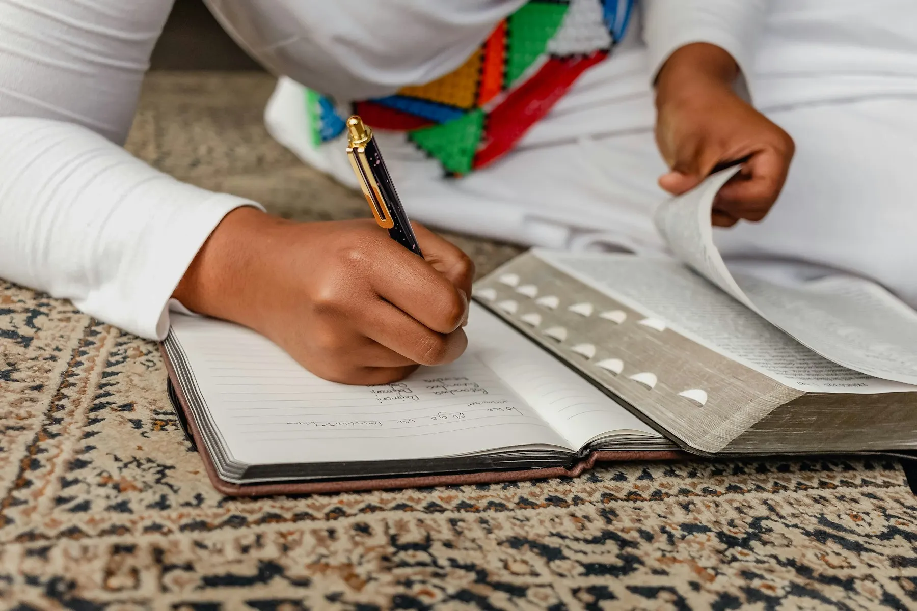 Las manos de una mujer con hiyab escriben en un cuaderno sobre una alfombra.