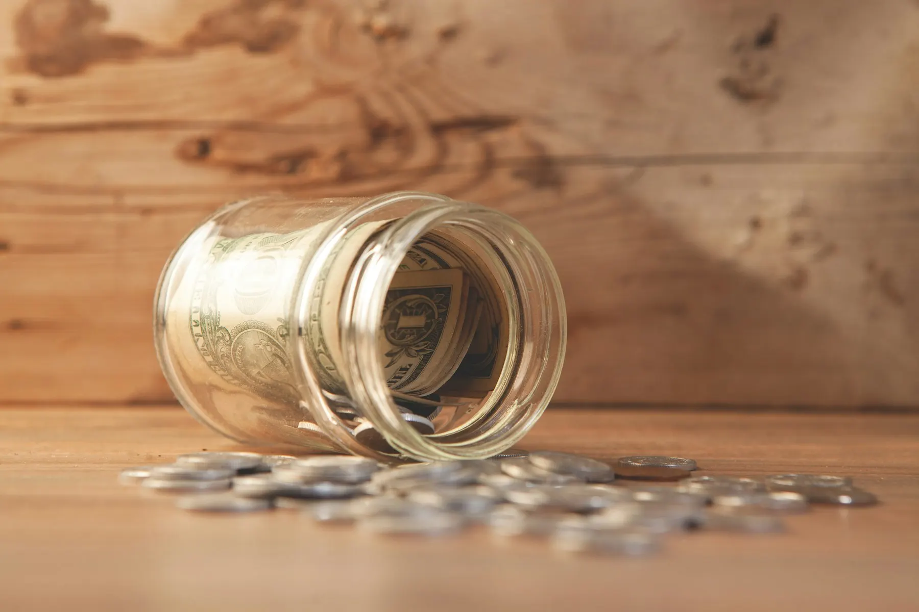 Un frasco de vidrio volcado con billetes y monedas derramados sobre una mesa de madera.