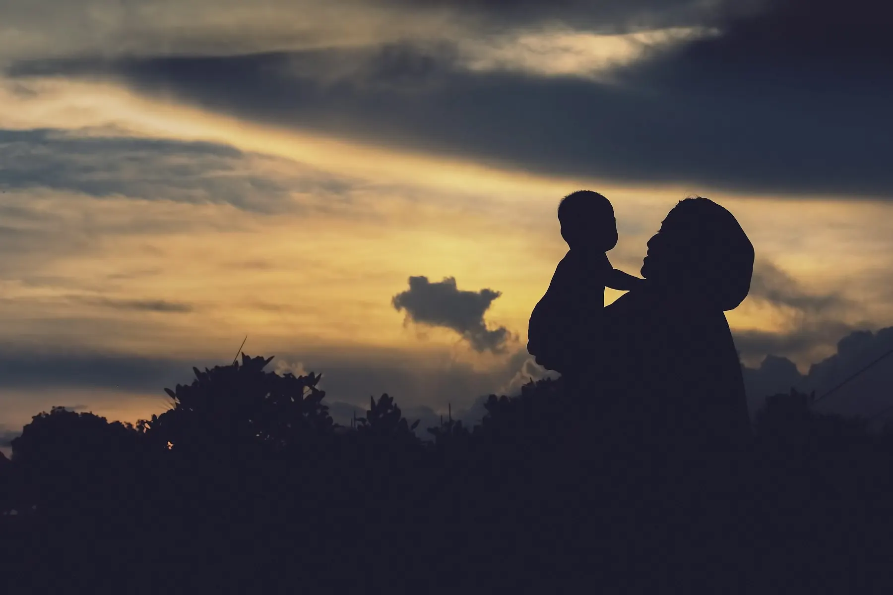 La silueta de una madre cargando a su hijo pequeño contra un cielo de atardecer, sin rostros visibles.