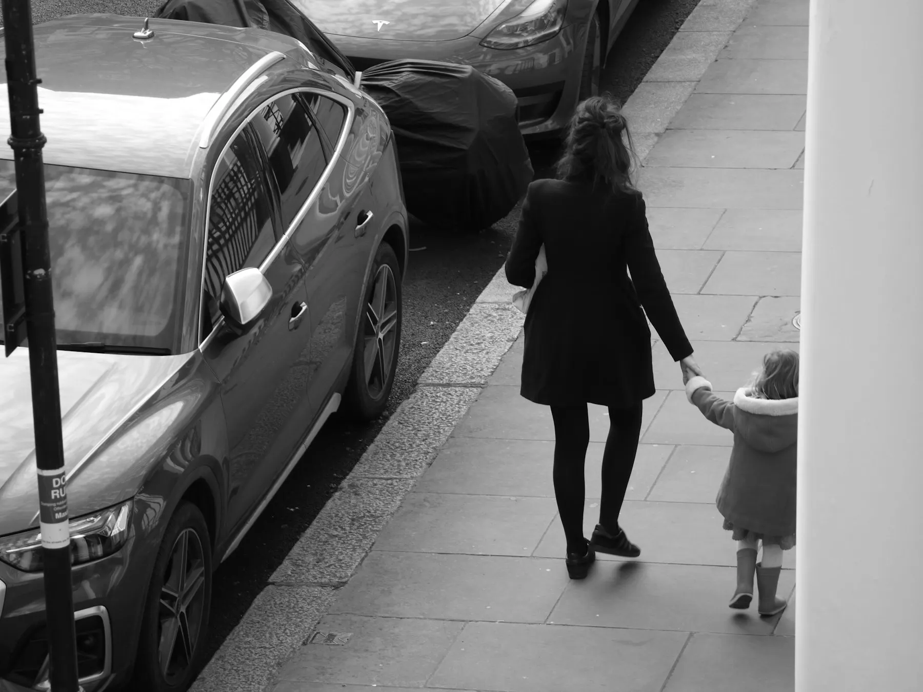 Una mujer camina de la mano de una niña por una banqueta, vistas de espaldas.